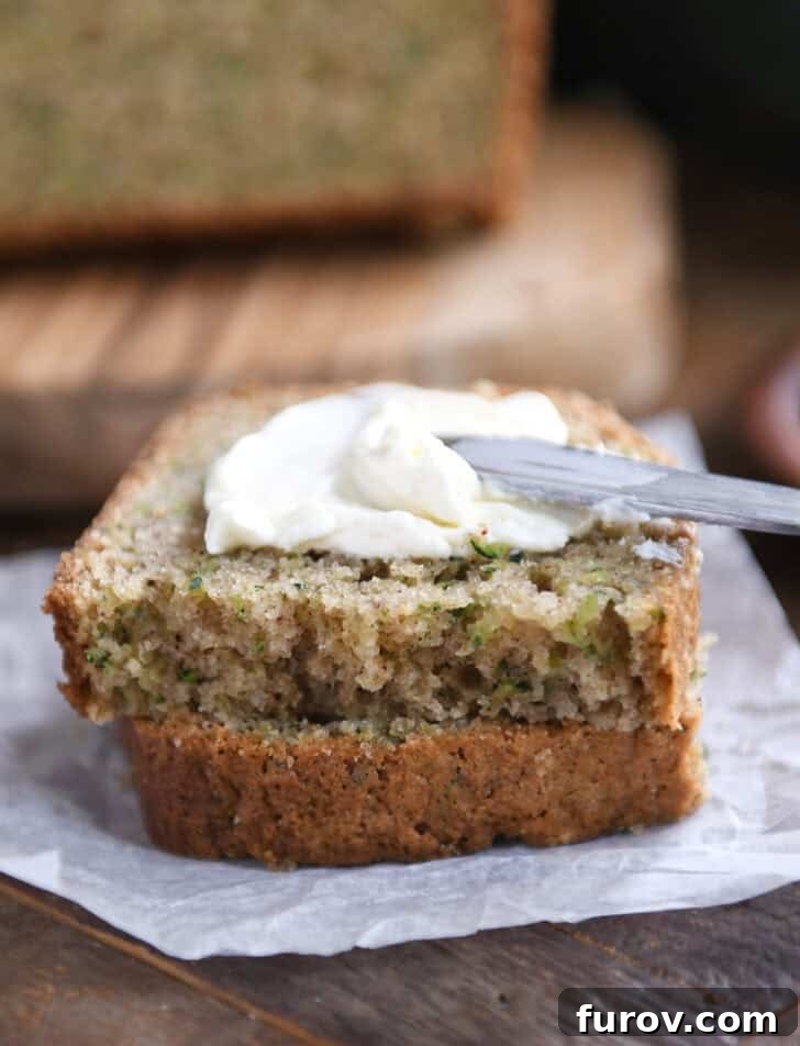 A hand gently spreading a generous layer of creamy, white cream cheese on top of a half-slice of warm zucchini bread, highlighting the lush texture and inviting appeal.