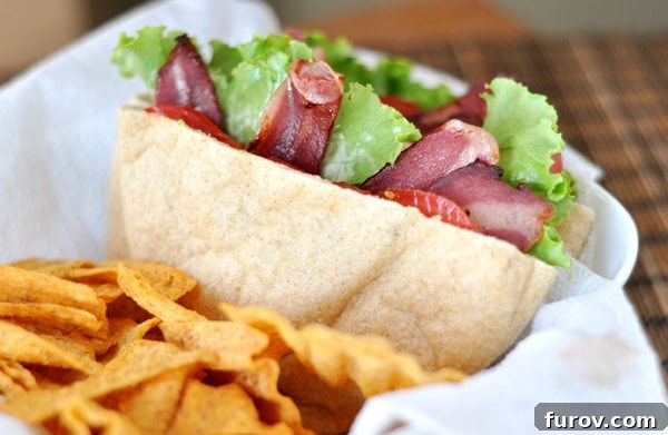 A close-up shot of a half-eaten BLT pita pocket, revealing the vibrant layers of turkey bacon, lettuce, tomato, and a creamy avocado spread, presented in a paper-lined basket with a side of chips.
