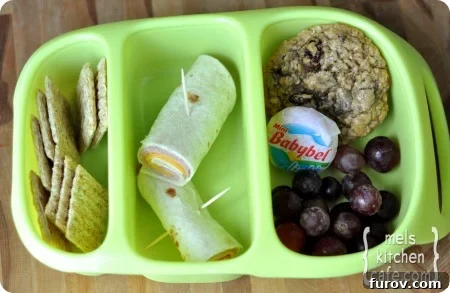 top view of a green divided lunch tray with crackers, ham cheese roll, grapes, Babybel, and cookie