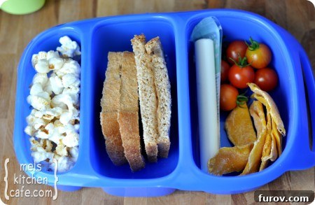 top view of a blue divided lunch box with popcorn, sandwich, cheese stick, tomatoes, and chips