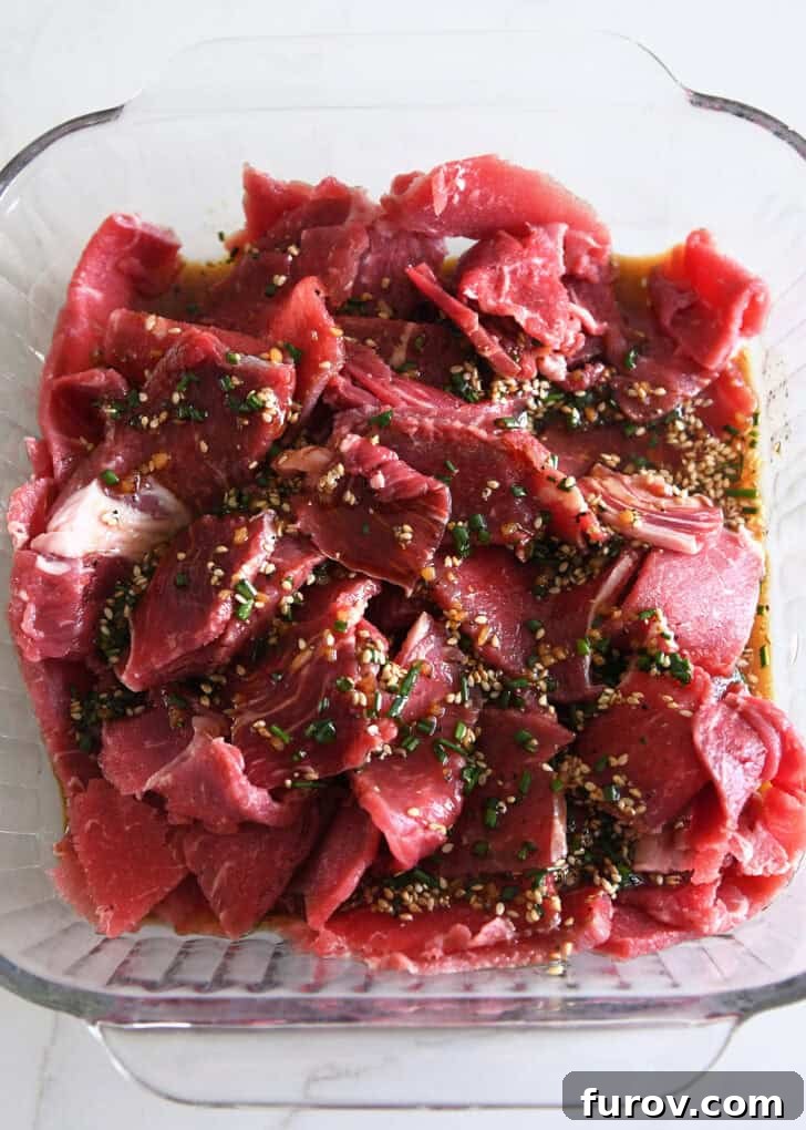 Top-down view of a square baking dish filled with thinly sliced beef submerged in a rich, savory marinade, garnished with fresh green onions and sesame seeds, ready for refrigeration.