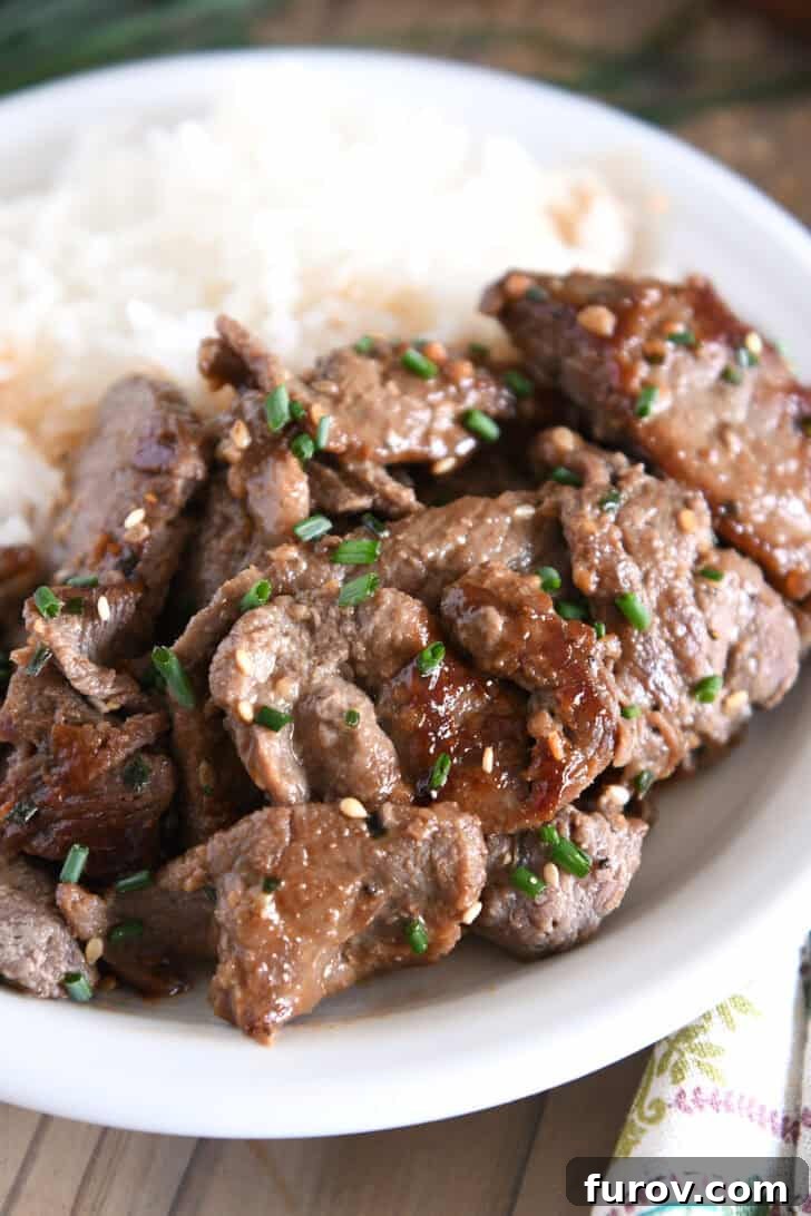 A beautifully presented white plate featuring a mound of fluffy white rice alongside several generous pieces of Korean beef bulgogi, garnished with vibrant green onions and toasted sesame seeds.
