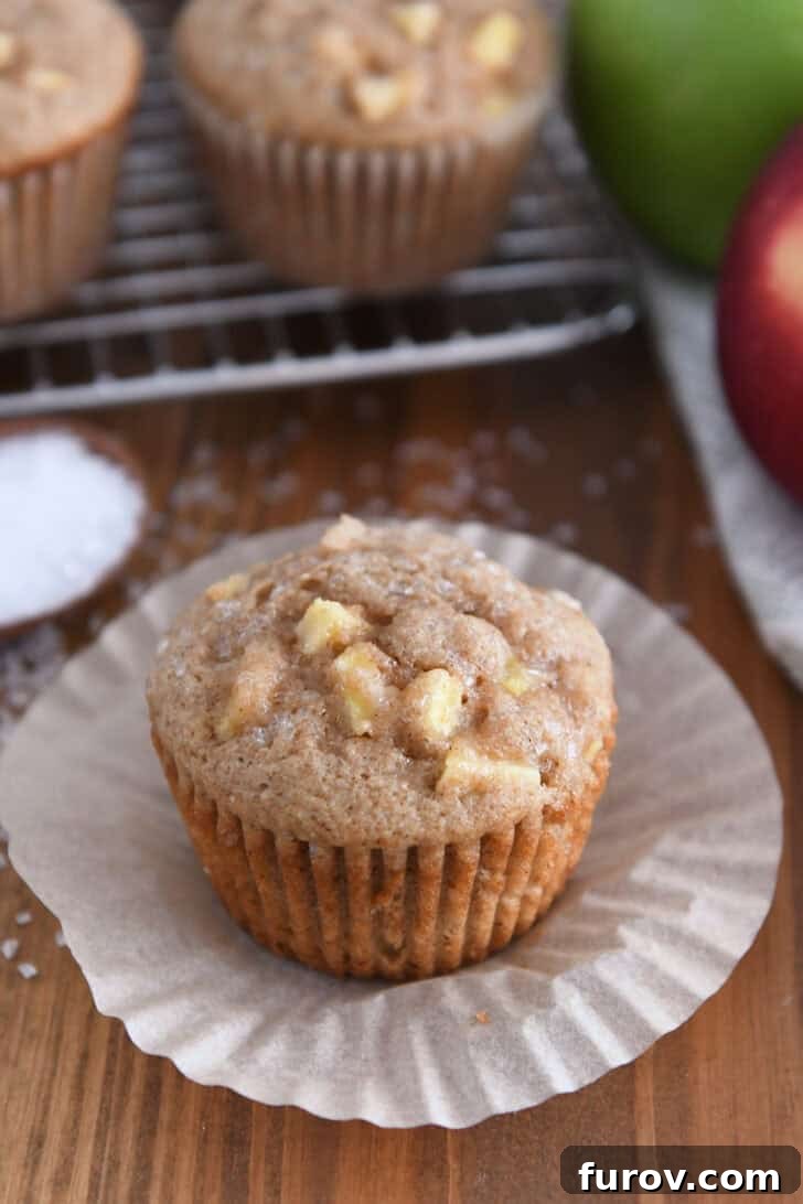 Spiced Apple Cinnamon Treats 10 An unwrapped apple muffin with a coarse sugar sprinkle in the background, surrounded by fresh apples, highlighting its rustic charm.