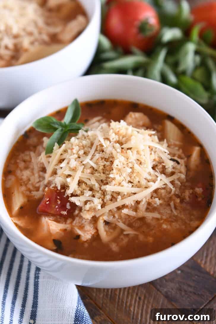 A captivating bowl of chicken parmesan soup, featuring perfectly cooked pasta, tender chicken, rich tomatoes, and fragrant seasonings, magnificently topped with golden buttered bread crumbs and freshly grated Parmesan cheese.