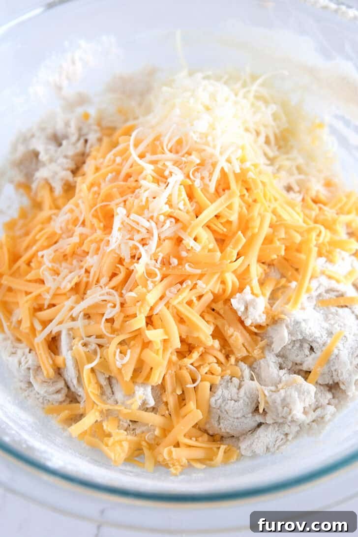 Sharp cheddar and Parmesan cheese sprinkled over shaggy bread dough in a glass bowl, ready to be incorporated.