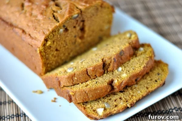 A perfectly baked loaf of pumpkin hazelnut bread, with several slices already cut and arranged on a white rectangular platter. A few whole hazelnuts and a sprig of fresh rosemary garnish the plate, emphasizing the autumnal theme.