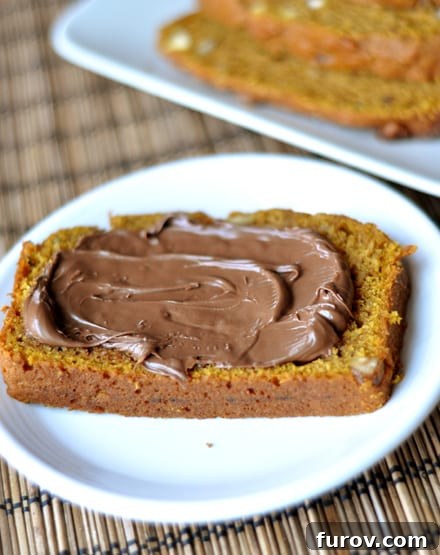 A thick, inviting slice of pumpkin bread generously slathered with rich, creamy Nutella, presented on a simple white plate. The focus is on the luxurious chocolate-hazelnut spread.
