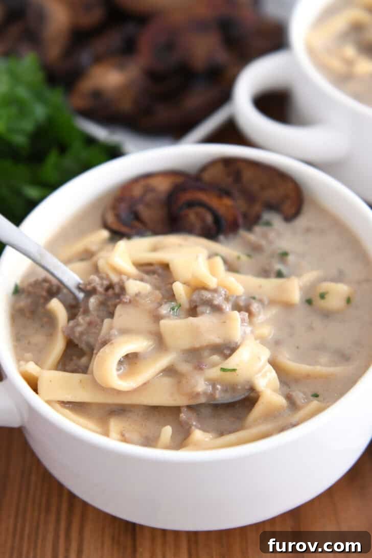 Spoon scooping up bite of creamy soup with noodles, ground beef, broth and mushrooms, garnished with parsley.