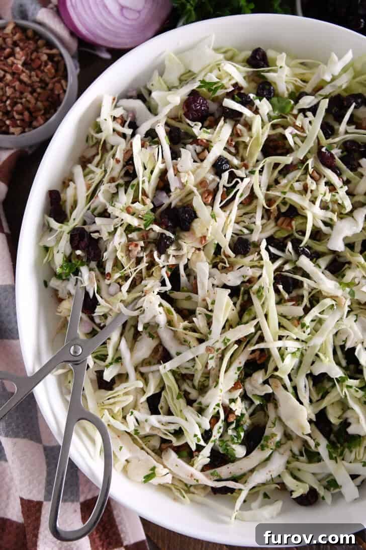 White bowl with salad tongs scooping portion of thin sliced green cabbage, dried cranberries, pecans and red onions.