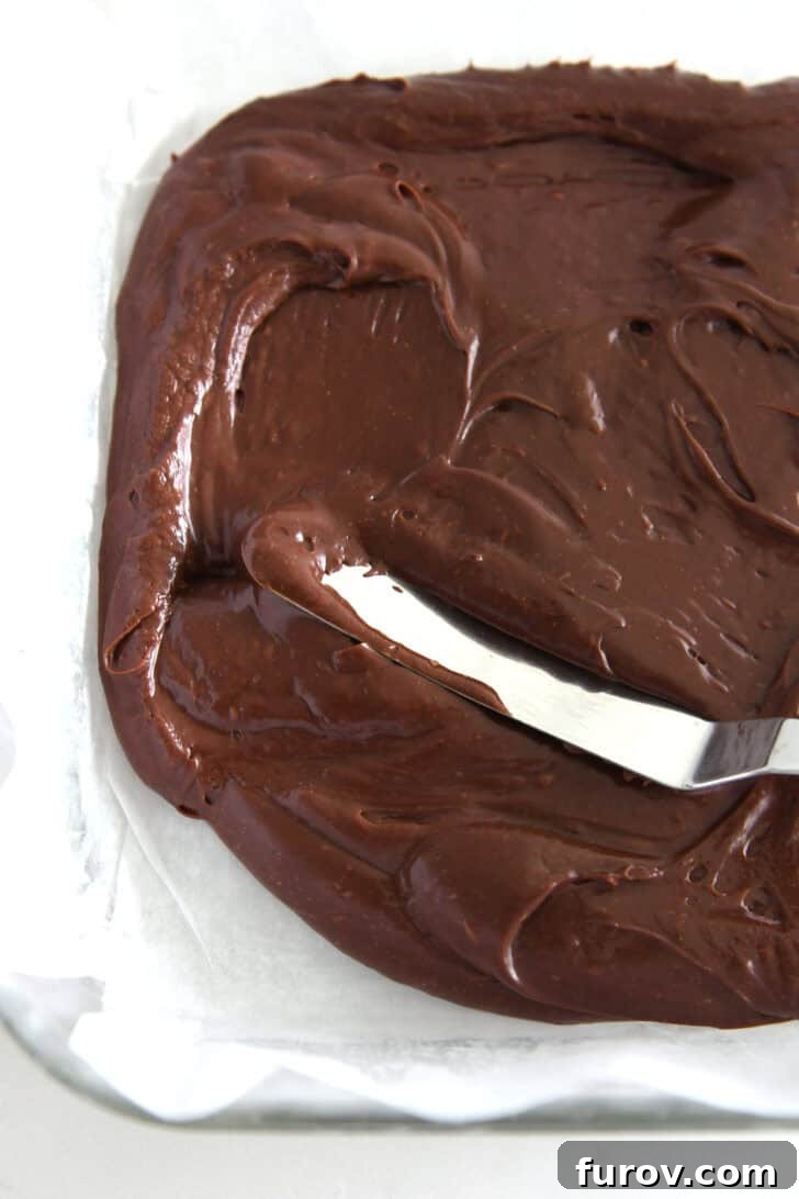Candy Cane Swirl Fudge 5 Rich, melted chocolate fudge being smoothly spread into a parchment-lined baking pan, creating the foundational layer for the peppermint bark fudge.