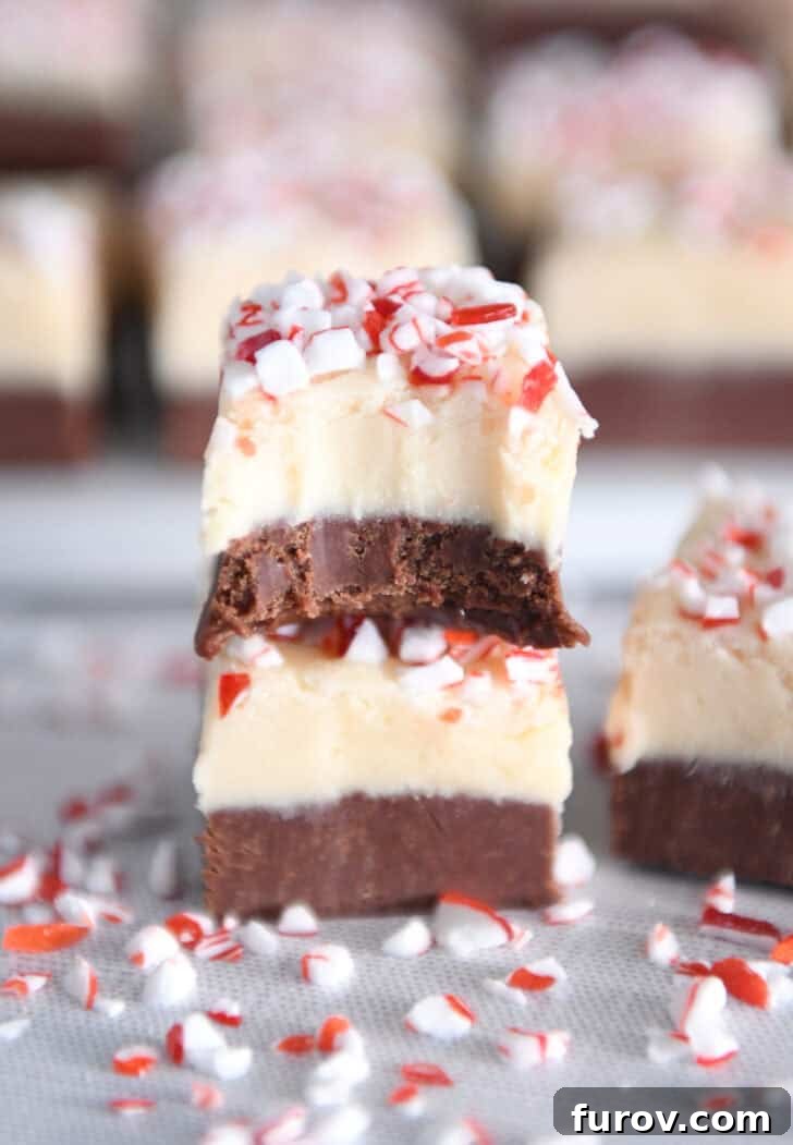Candy Cane Swirl Fudge 8 Two pieces of layered peppermint bark fudge, one stacked on top of the other, on a simple white background. The top piece has a perfect bite taken out, revealing the distinct dark and white chocolate layers.