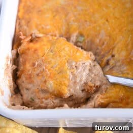 Serving spoon scooping serving of warm bean dip topped with cheese in white square baking dish.