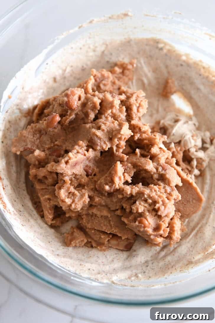 A medium glass bowl showcasing refried beans being incorporated into the already creamy sour cream and spice mixture, demonstrating the beginning of the blend.