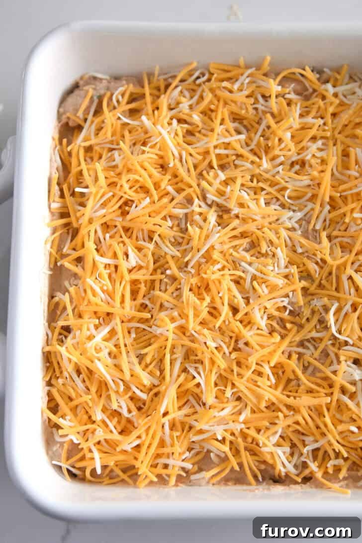 The white square baking pan, now generously topped with shredded cheese over the bean dip, poised for baking to golden, bubbly perfection.