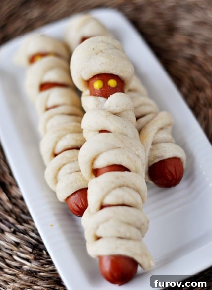 Hot dogs wrapped in breadstick dough to look like mummies on a white platter, with ketchup and mustard eyes.