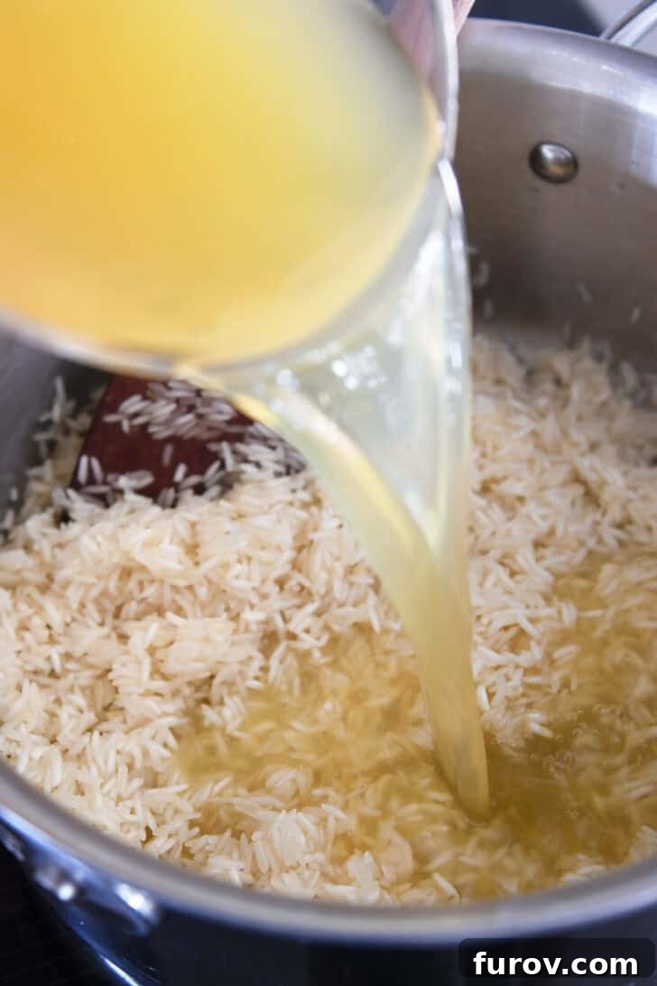 A steady stream of rich chicken broth being poured into a pan containing butter-coated rice, ready for cooking.