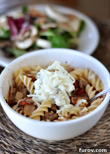 A beautifully presented white bowl of lasagna soup, garnished with a dollop of creamy ricotta mixture and fresh herbs, with a vibrant green salad subtly blurred in the background, suggesting a complete and satisfying meal.