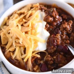 White bowl with handles and spoon scooping bite of beef and bean chili with shredded cheese, sour cream and corn chips.