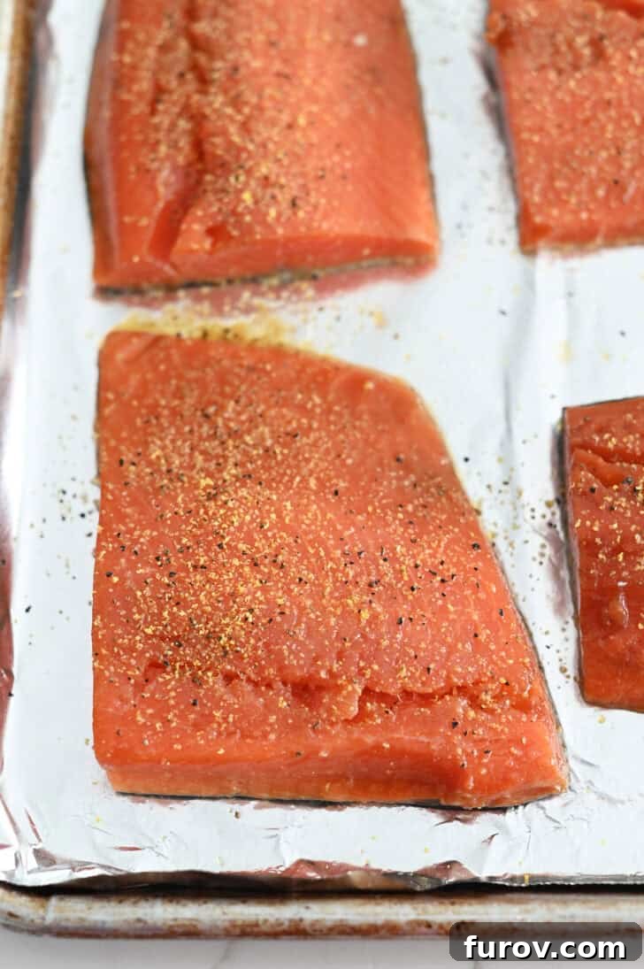 Salmon fillets generously sprinkled with vibrant lemon pepper seasoning, arranged on a foil-lined baking sheet, prepped for baking.