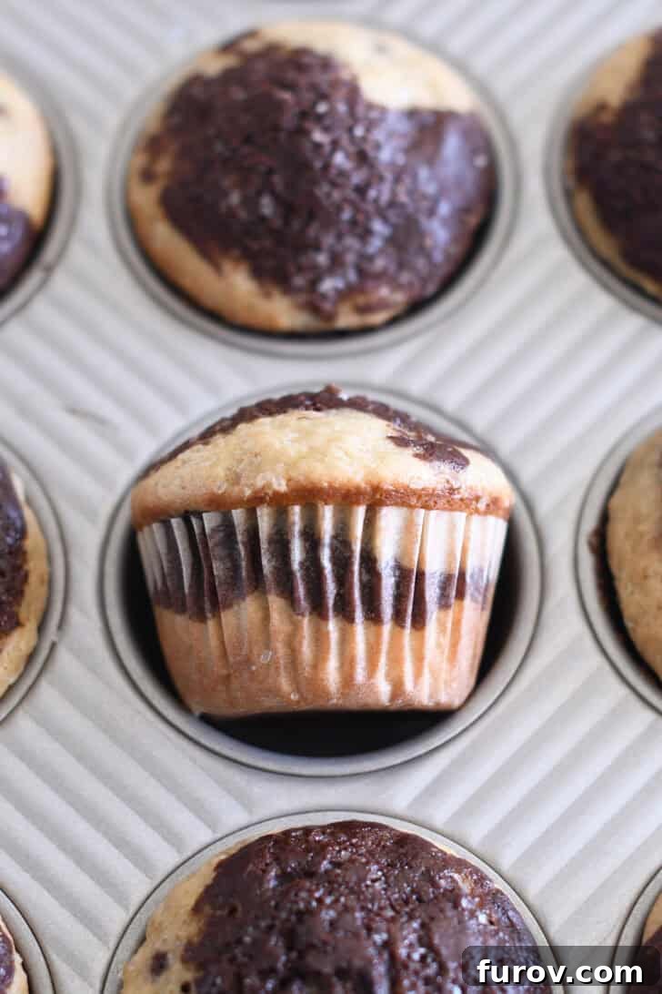 Marbled Chocolate Banana Muffins 12 A delightful chocolate and banana muffin, turned on its side within its muffin tin liner, revealing the beautiful swirl pattern and golden-brown exterior.