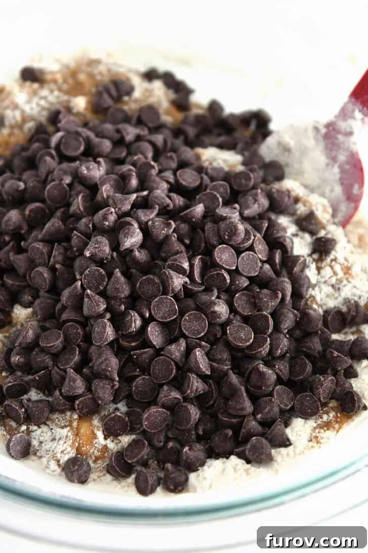 Semisweet chocolate chips in a glass bowl alongside dry ingredients and a red silicone spatula, ready to be incorporated into the dough.