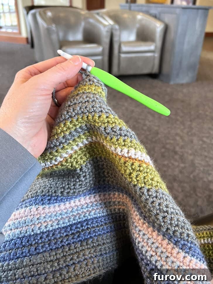 Close-up shot of hands crocheting a gray row on a multi-colored temperature blanket, possibly in a doctor's office setting.