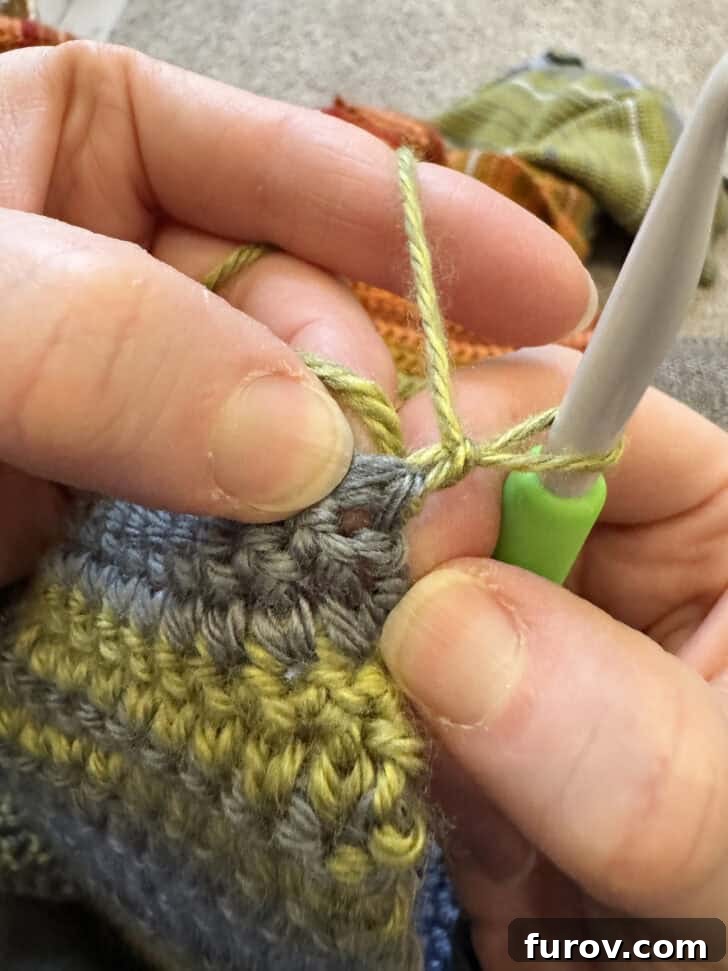 Close-up showing a crochet hook inserting into the first stitch of the second row, demonstrating the correct placement after turning the blanket.