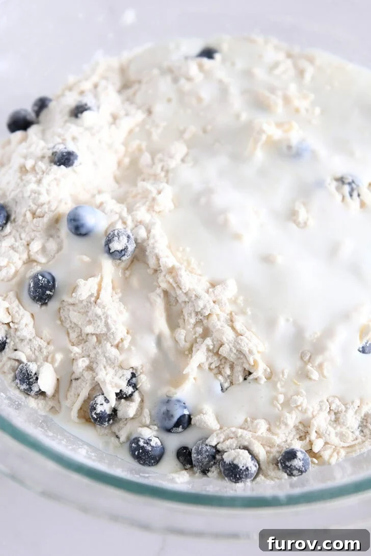 Buttermilk and fresh lemon juice being poured into a glass bowl containing scone dough and blueberries, illustrating the gentle mixing stage.