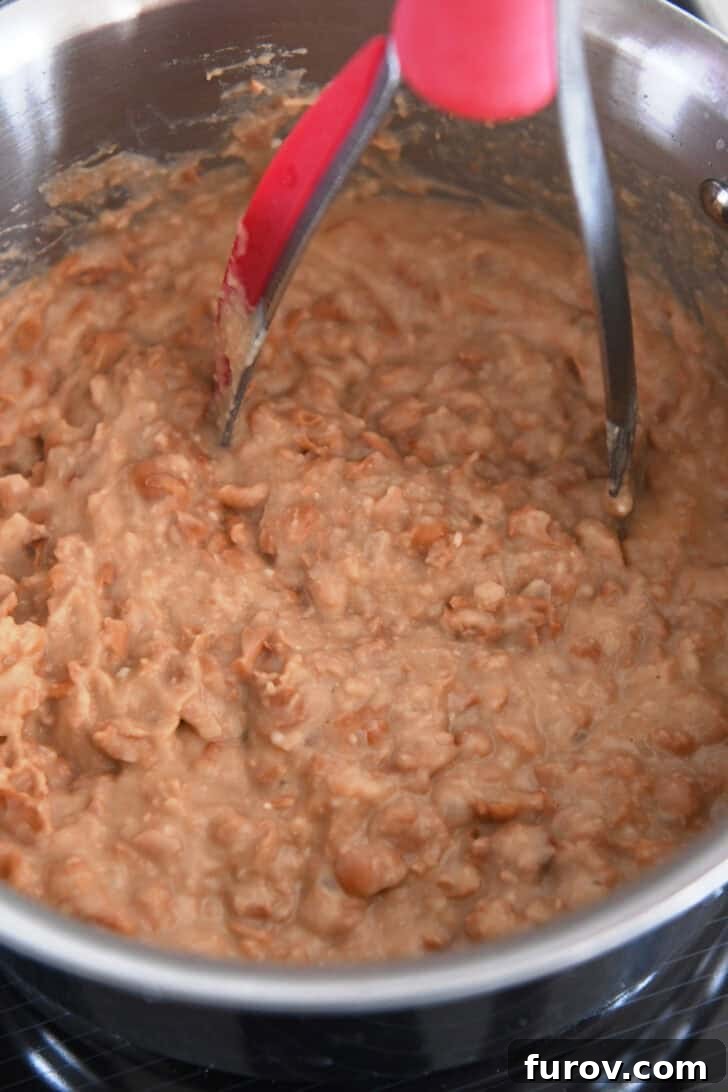 A sturdy potato masher actively mashing soft canned pinto beans directly in the stainless steel pot, achieving the desired refried bean texture.