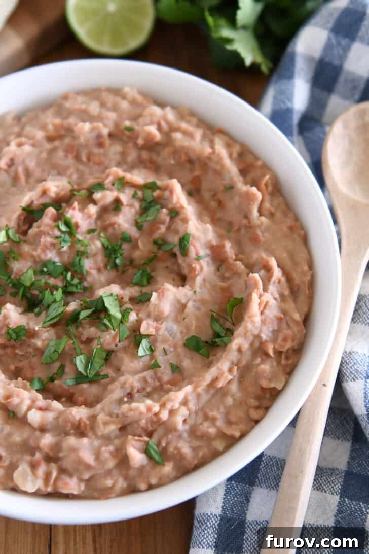 A pristine white bowl filled with easy creamy refried beans, elegantly garnished with a sprig of fresh cilantro, highlighting their appetizing appearance.