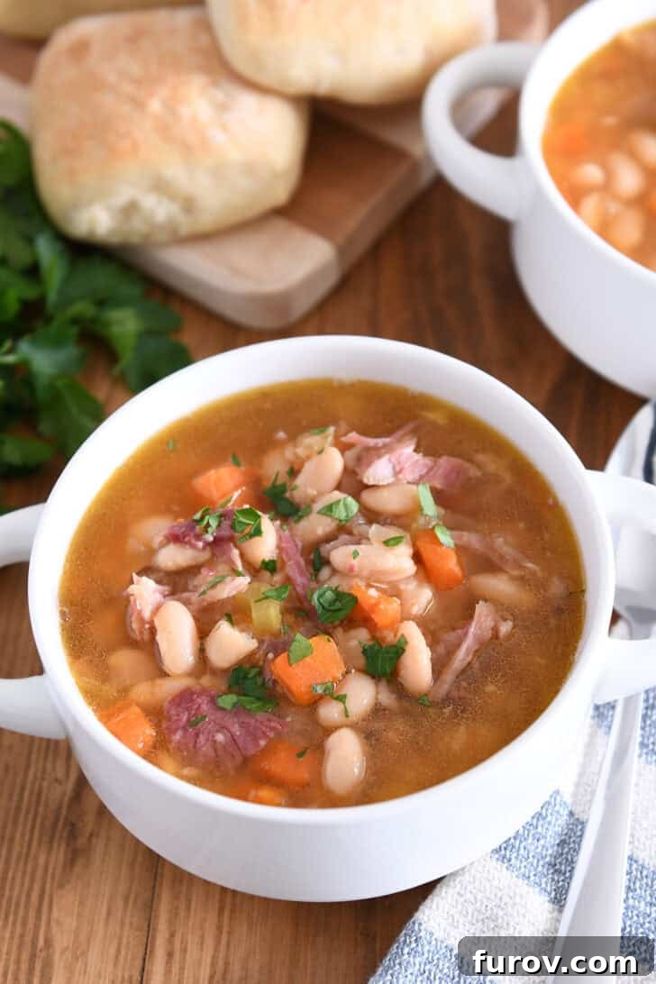 A rustic white bowl with handles filled to the brim with slow cooker ham and bean soup, garnished with fresh herbs.
