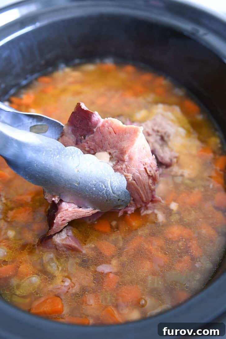 Tongs lifting a meaty ham bone out of the slow cooker, revealing tender white beans, broth, and vegetables.