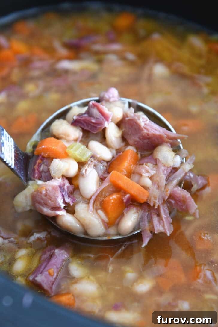 A ladle holding a generous scoop of slow cooker ham and bean soup, showing tender beans, carrots, celery, and ham pieces.