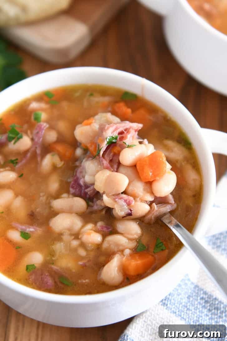 Top-down view of a white bowl with handles, showcasing a spoon lifting a perfect bite of slow cooker ham and bean soup with white beans, carrots, and ham.