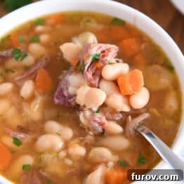White bowl with handles, with a spoon lifting a bite of white beans, carrots, and ham from the slow cooker soup.