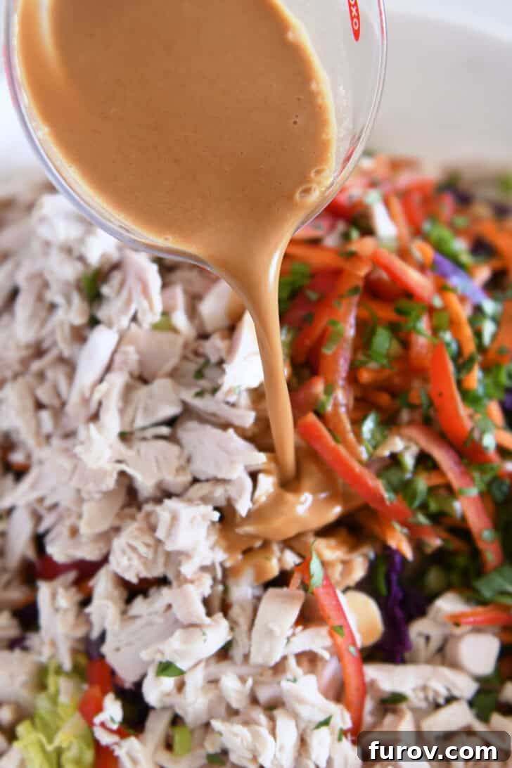 Pouring homemade peanut vinaigrette dressing over a vibrant mix of Asian chicken salad ingredients in a white bowl.