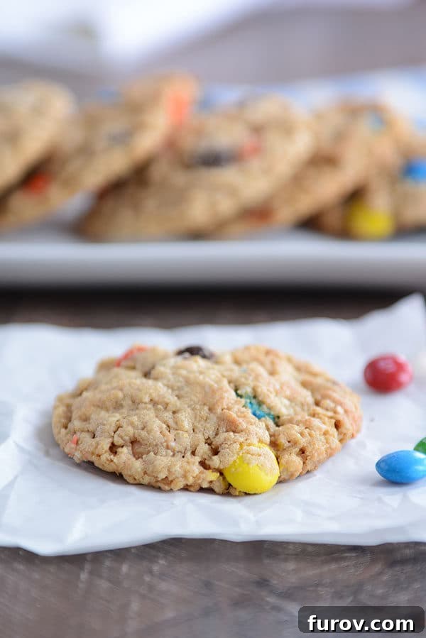 Epic One-Bowl Gluten-Free M&M Monster Cookies 3 An M&M oat cookie cooling on a piece of parchment paper, with a baking tray full of more cookies in the soft background.