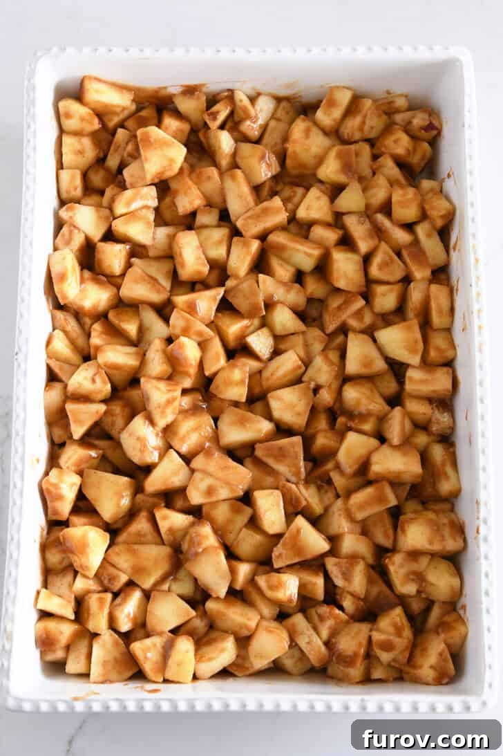 Gooey Caramel Apple Crumble 5 Top-down view of cinnamon-spiced chopped apples generously mixed with caramel sauce in a white 9x13-inch baking dish, ready for the topping.