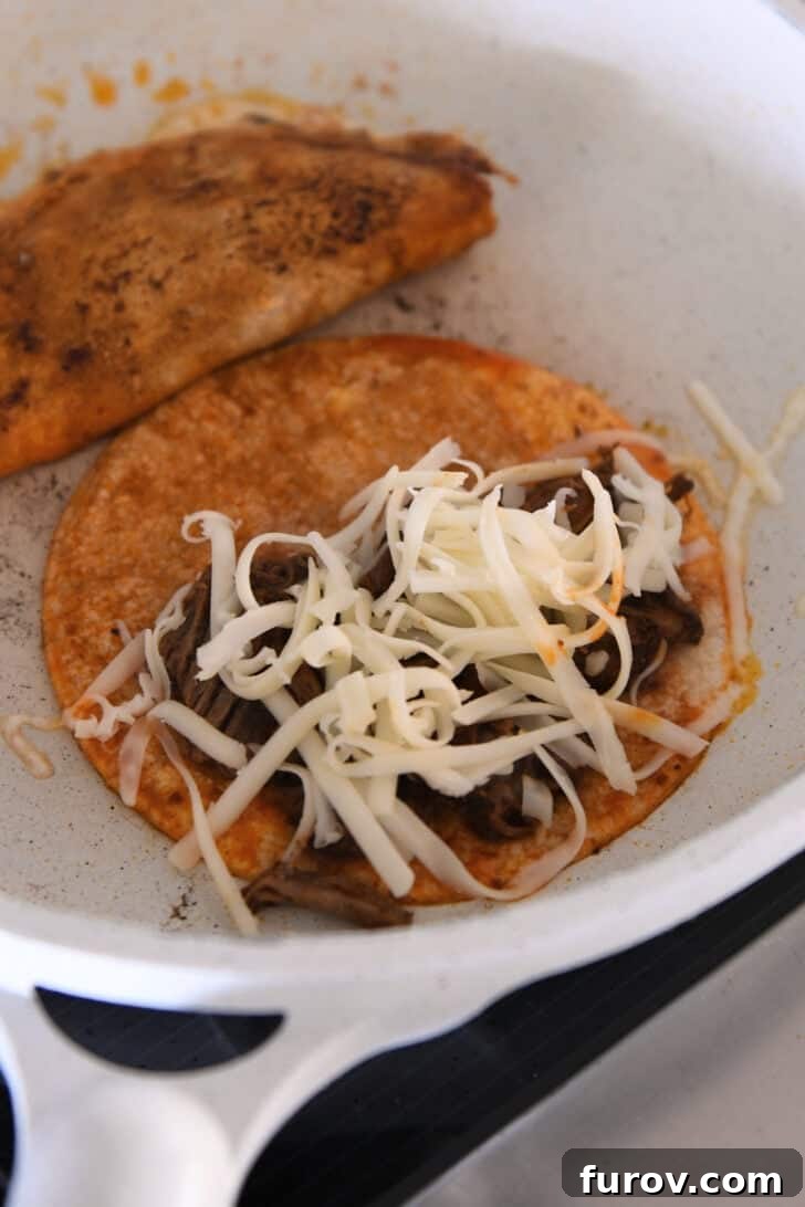 Ultimate Slow Cooker Birria Tacos 9 White skillet with two corn tortillas soaked in birria broth topped with shredded beef and shredded cheese.