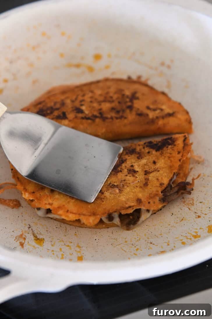 Ultimate Slow Cooker Birria Tacos 10 Pressing crispy corn tortilla taco in white skillet with metal spatula.