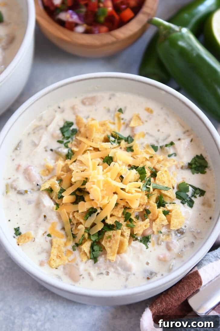 Velvety White Chicken Chili 2 Top down view of a white bowl of white chicken chili topped with shredded cheddar cheese, chopped cilantro, and crushed tortilla chips, ready to be served.