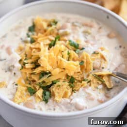 Velvety White Chicken Chili 7 A white bowl of white chicken chili topped with shredded cheddar cheese, chopped cilantro, and crushed tortilla chips, a perfect and easy meal.