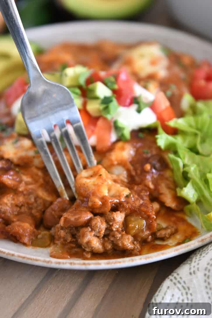 Fork scooping a bite of ground beef enchiladas with red sauce on a gray plate, garnished with fresh lettuce, sour cream, avocado slices, and cilantro.