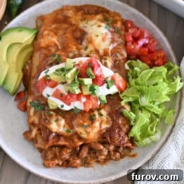 Top down view of two enchiladas on gray plate topped with red enchilada sauce, cheese, cilantro, sour cream, tomatoes, and avocados.