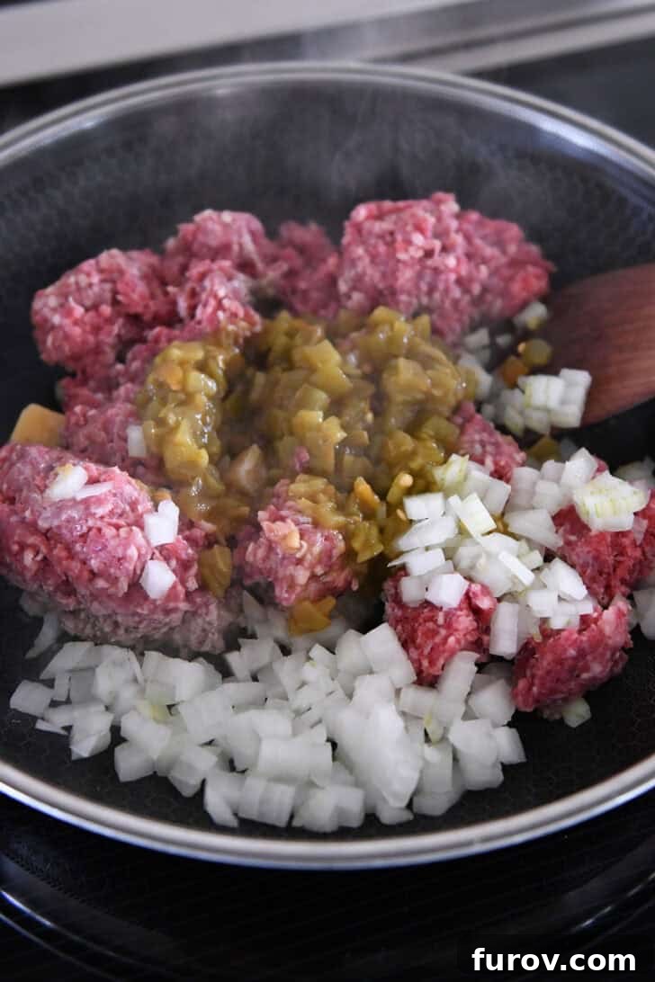 Ground beef, finely chopped onions, and green chiles cooking in a 12-inch nonstick skillet on an induction stovetop, browning evenly.