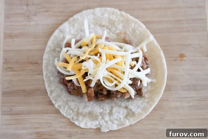 Top-down view of a 6-inch flour tortilla, meticulously topped with a generous portion of ground beef enchilada filling and a sprinkle of shredded cheddar and Monterey Jack cheese, ready for rolling.
