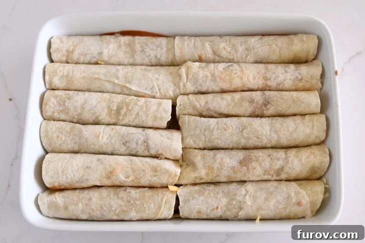 Ten perfectly rolled ground beef enchiladas nestled in flour tortillas, arranged in two rows in a white 9x13-inch baking dish, ready for sauce and cheese.