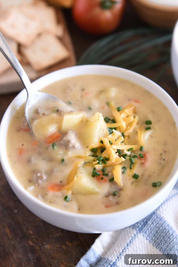 A white bowl with a spoon scooping creamy cheesy soup. The soup contains cooked potatoes, ground beef, and carrots, and is topped with shredded cheddar cheese and fresh chopped chives. This delightful dish is a lightened up cheeseburger soup from melskitchencafe.com.