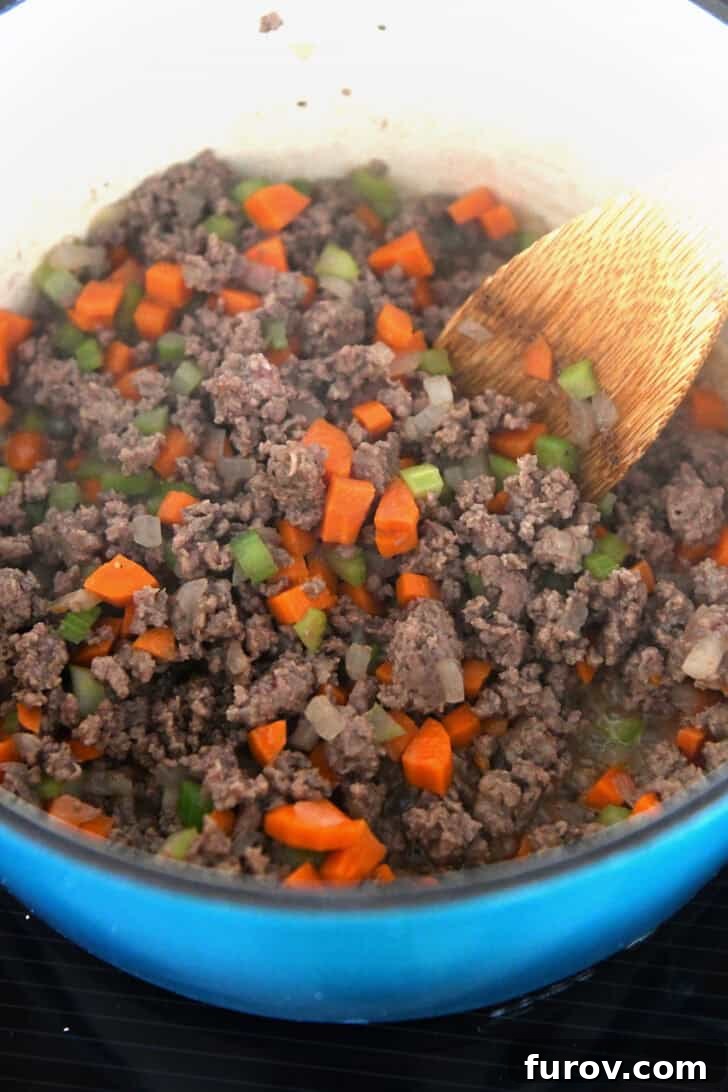 An enameled cast iron pot contains cooked ground beef, diced carrots, and celery, ready for the next steps in preparing the lightened up cheeseburger soup.