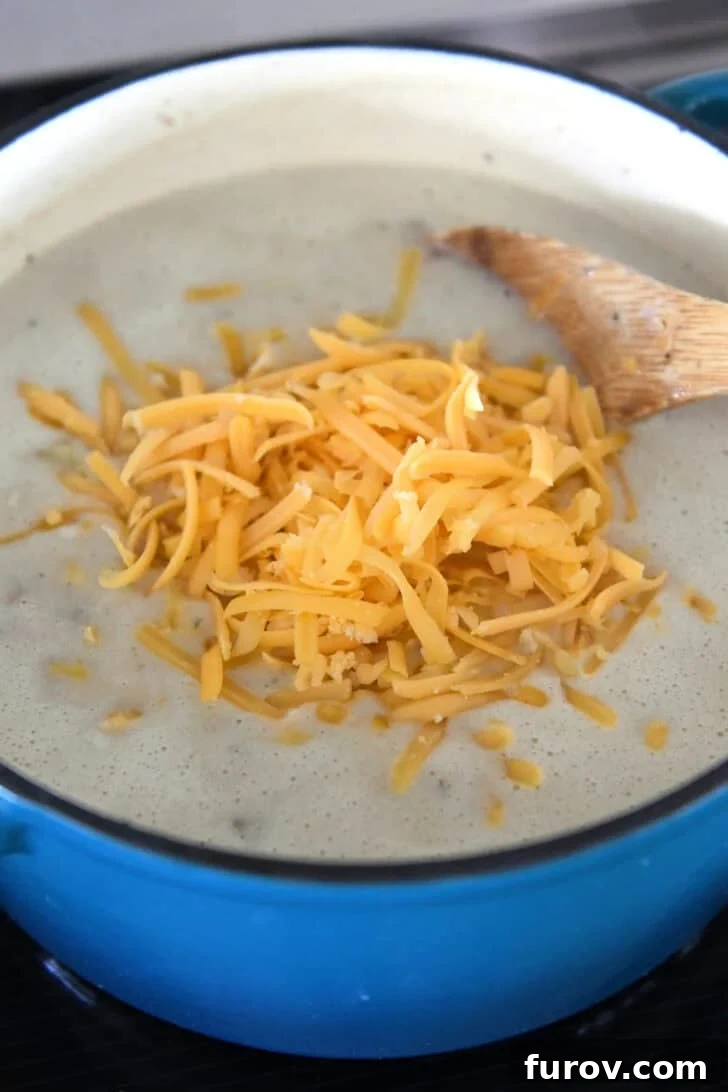 An enameled cast iron Dutch oven holds a creamy soup, with a generous pile of shredded sharp cheddar cheese in the center, about to be stirred in.